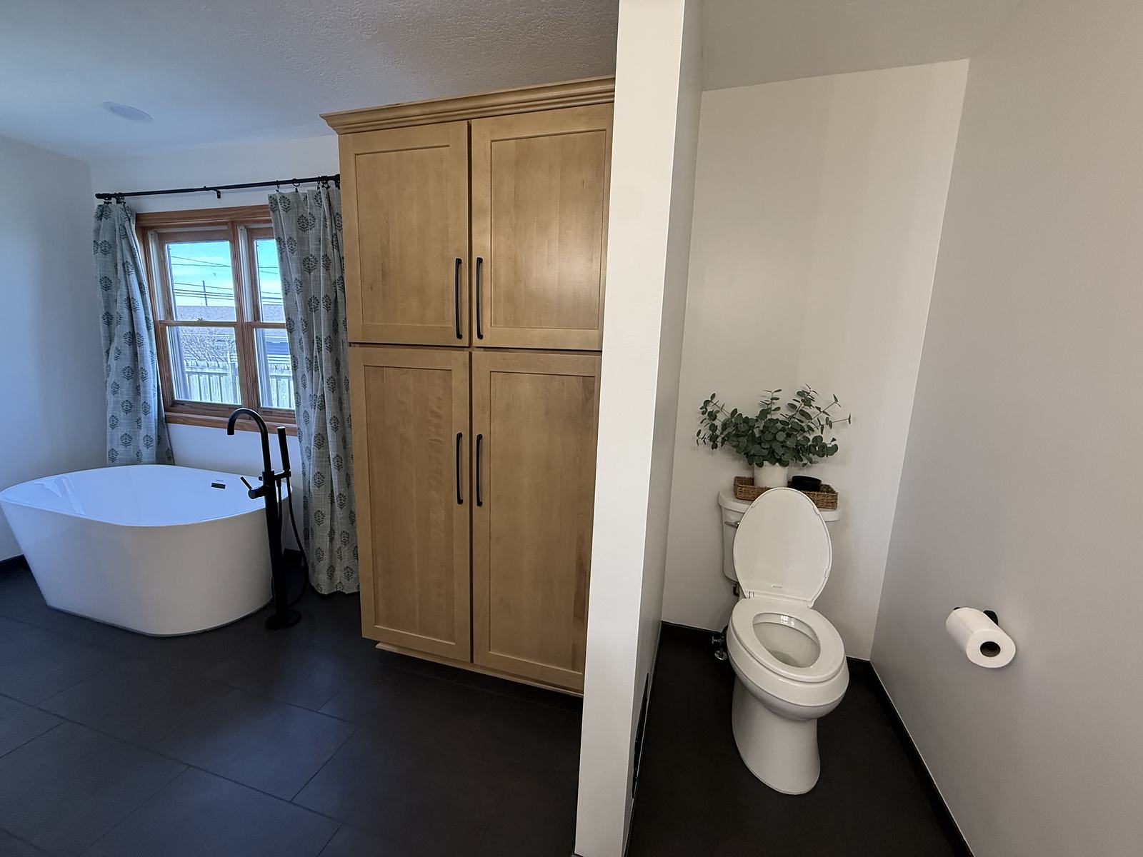 Freestanding soaking tub beside a maple linen tower and a private toilet nook