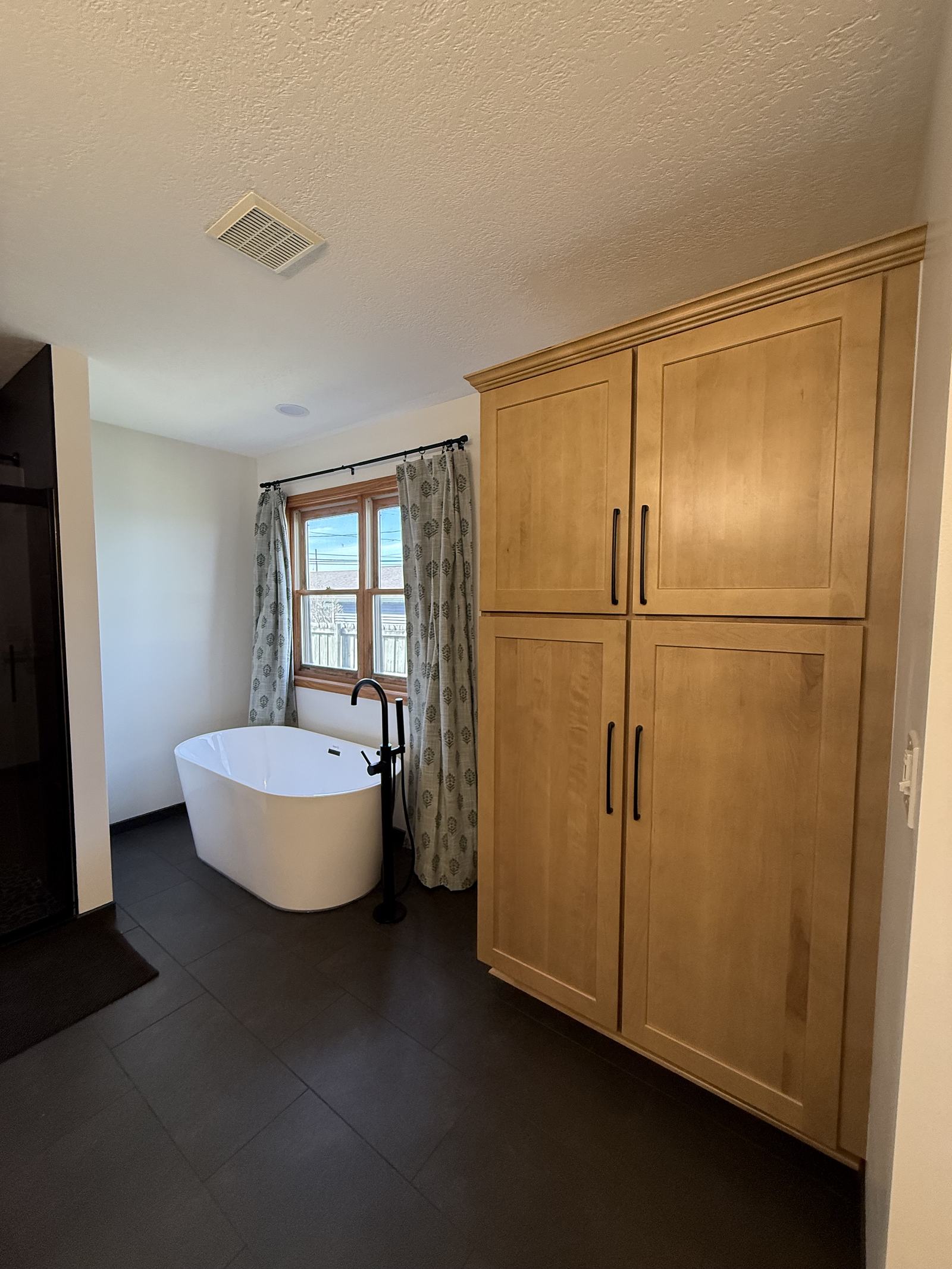 Maple floor-to-ceiling linen tower next to a freestanding tub on charcoal tile